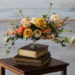Low arrangement of yellow and peach roses and ranunculus in a silver pedestal bowl on stacked books.