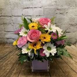 Mixed bouquet of pink roses, daisies, and yellow flowers in a square vase