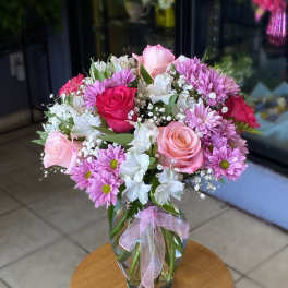 Pink roses, lavender daisies, and white blooms in a clear glass vase with a pink ribbon.