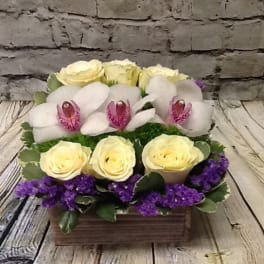 Arrangement of pale yellow roses, white orchids, and purple filler in a wooden box