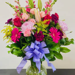 Mixed bouquet of pink, purple, and yellow flowers in a glass vase with a lavender ribbon.