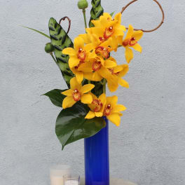 Yellow orchids arranged in a tall blue vase with candles nearby.