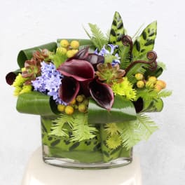 Tropical floral arrangement with dark calla lilies and purple blooms in a glass vase