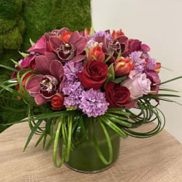 Low arrangement of red roses, burgundy orchids, and lavender flowers in a clear glass cylinder vase