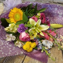 Mixed bouquet with a sunflower, lilies, roses, and purple blooms wrapped in purple paper