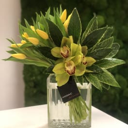 Yellow tulips and green orchids arranged with lush foliage in a clear glass vase