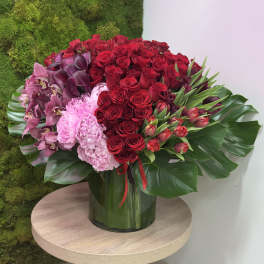 Large arrangement of red roses, pink peonies, orchids and tulips in a glass vase with broad tropical leaves.