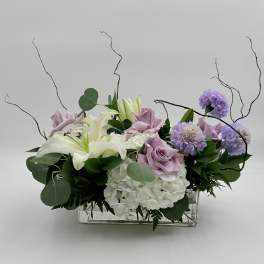 Lavender roses and white lilies arranged with purple blooms in a clear rectangular vase.