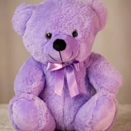 Lavender plush teddy bear with a satin bow sitting against a neutral background.