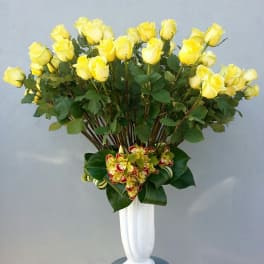 Tall arrangement of yellow roses in a white vase with a small orchid cluster at the base
