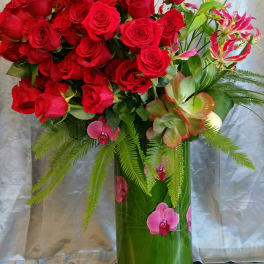 Red roses arranged in a tall glass vase with pink orchids and green foliage.