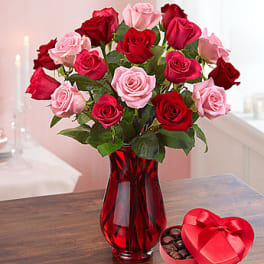 Bouquet of red and pink roses in a red glass vase beside a heart-shaped chocolate box.
