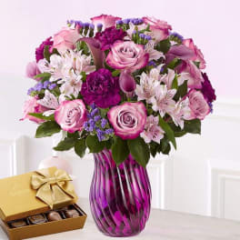 Pink and purple bouquet in a glass vase beside a box of chocolates