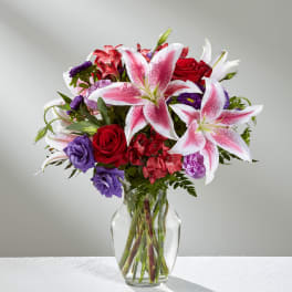 Bouquet of pink lilies, red roses, and purple flowers in a clear glass vase