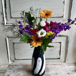 Mixed bouquet with orange gerberas, purple flowers, and roses in a black vase