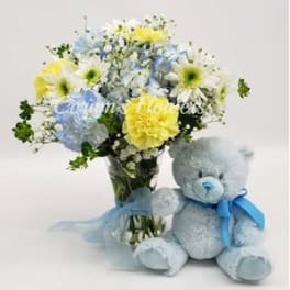 Bouquet of blue, yellow, and white flowers in a glass vase beside a blue teddy bear