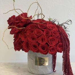 Red rose arrangement in a gray cylindrical box with trailing burgundy accents