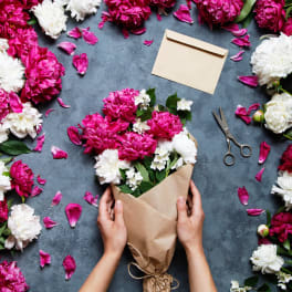 Hand-held bouquet of pink and white peonies in brown paper with an envelope and scissors nearby