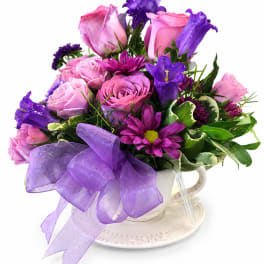 Pink and purple flower arrangement in a white teacup with a lavender bow