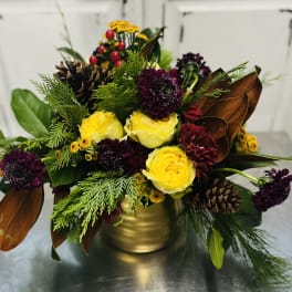 Yellow and burgundy flowers arranged in a gold vase with pinecones and greenery.