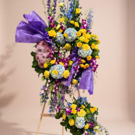 Tall floral standing arrangement with yellow roses, blue hydrangeas, and purple orchids on an easel