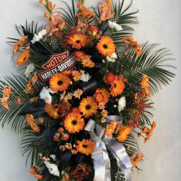 Tall standing spray with orange gerbera daisies, white flowers, palm fronds, and a Harley-Davidson logo sign.