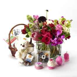 Flower arrangement in a glass vase with a gift basket, teddy bear, chocolates, and bath items