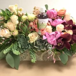Mixed floral arrangement with roses, orchids, succulents, and a bath oil bottle in a decorative box
