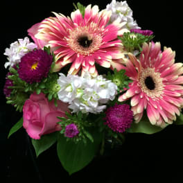 Pink gerbera daisies, roses, and white blossoms arranged in a small vase against a black background