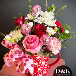 Pink and red flower arrangement in a heart-covered mug with a ribbon bow