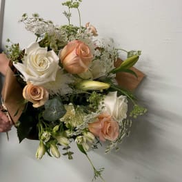 Hand-tied bouquet of white and peach roses with lilies and delicate white filler flowers in kraft paper wrap.