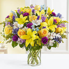 Bright mixed bouquet of yellow lilies, roses, and purple flowers in a glass vase