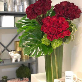 Tall arrangement of clustered red roses with large tropical green leaves in a clear glass vase