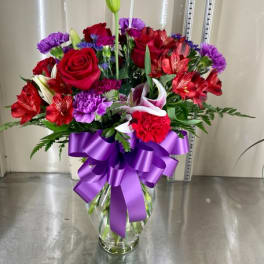 Bouquet of red roses, lilies, and purple flowers in a glass vase with a purple ribbon