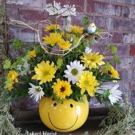 Yellow and white daisy arrangement in a smiley-face pot with a small bird decoration