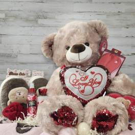 Large teddy bear holding a heart pillow with assorted gift items