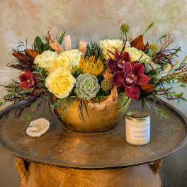 Arrangement of cream roses and burgundy orchids in a gold bowl with a candle
