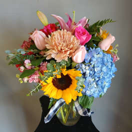 Mixed bouquet in a glass vase with sunflowers, pink roses, lilies, and blue hydrangeas