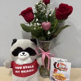 Three red roses in a glass vase beside a plush raccoon and gift card
