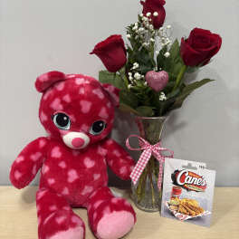 Red roses in a glass vase beside a pink heart-pattern teddy bear and gift card