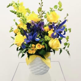 Yellow roses and blue irises in a white vase with a yellow ribbon