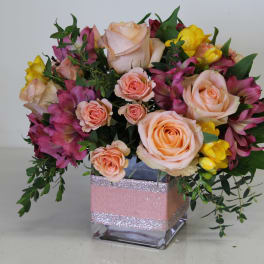 Short arrangement of peach roses, yellow freesia, and pink alstroemeria in a glass cube vase with pink ribbon.