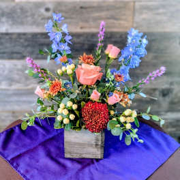 Mixed arrangement of pink roses, blue delphinium, and red mum in a wooden cube vase