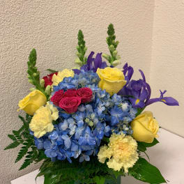 Bouquet of blue hydrangeas, yellow roses, and purple irises in a square vase
