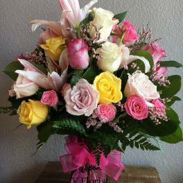 Tall mixed arrangement of pink lilies and pink, yellow, and white roses in a clear vase with pink bow