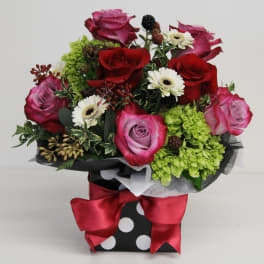 Compact arrangement of red and pink roses with white daisies in a polka dot box with red bow
