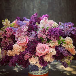 Pink roses in a decorative vase surrounded by purple and lavender blossoms