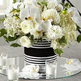 White floral arrangement in a striped black-and-white vase with candles around it