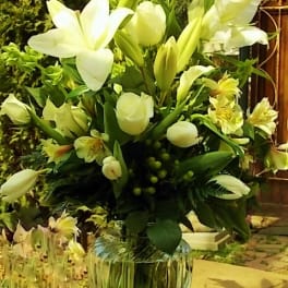 White lilies and pale yellow flowers arranged in a clear glass vase.