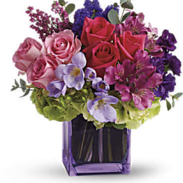 Compact arrangement of pink and purple flowers in a purple glass cube vase